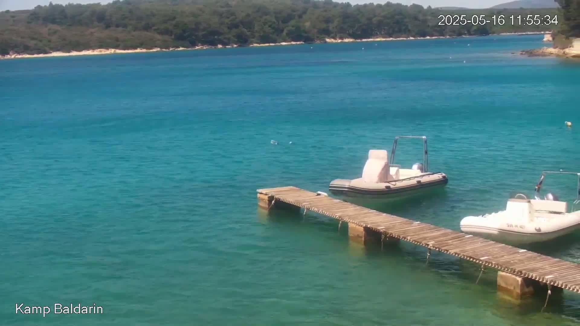 Two inflatable boats are moored at a wooden dock in a calm, clear turquoise bay, with a green-covered shoreline in the background.