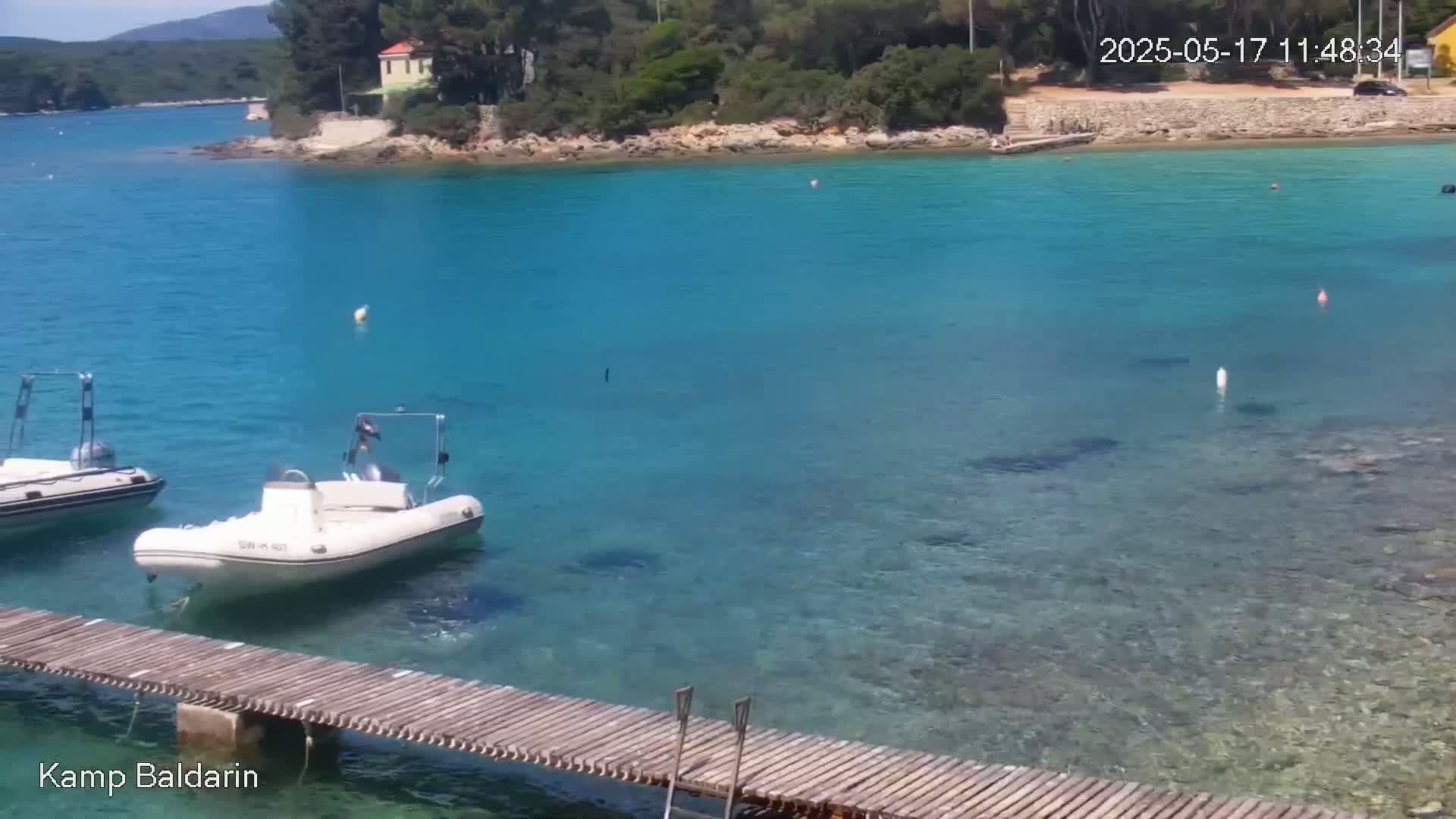 A wooden dock extends over clear, turquoise water near a small, rocky shoreline with some trees and a building, on a sunny day.
