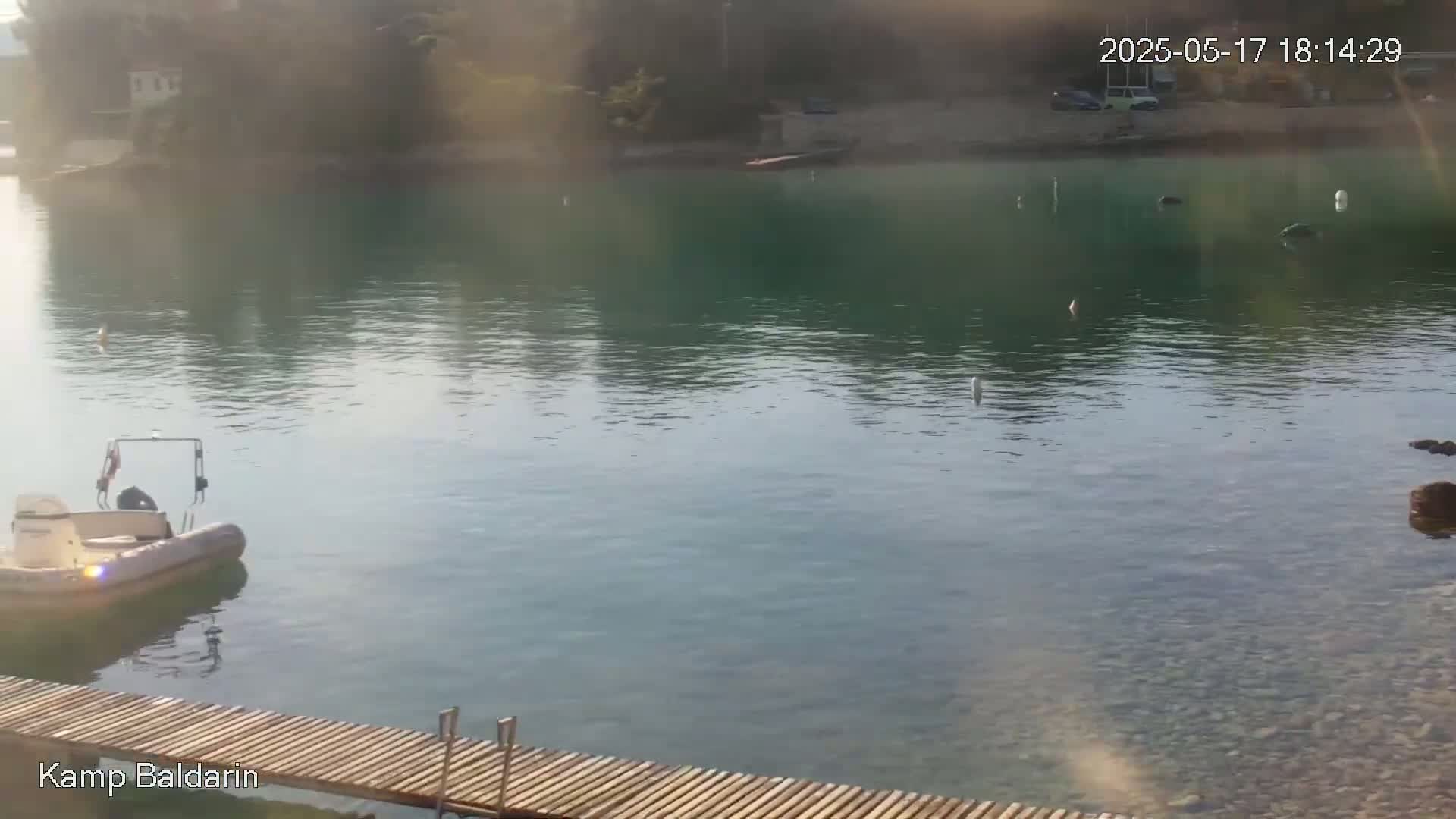 A small motorboat is docked near a wooden pier on calm, clear, teal water next to a rocky shoreline and a distant wooded area.
