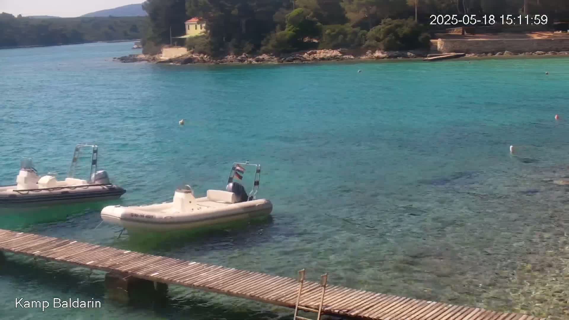 Two motorboats are moored near a wooden dock in a calm, clear turquoise bay, with green hills visible in the distance under a sunny sky.