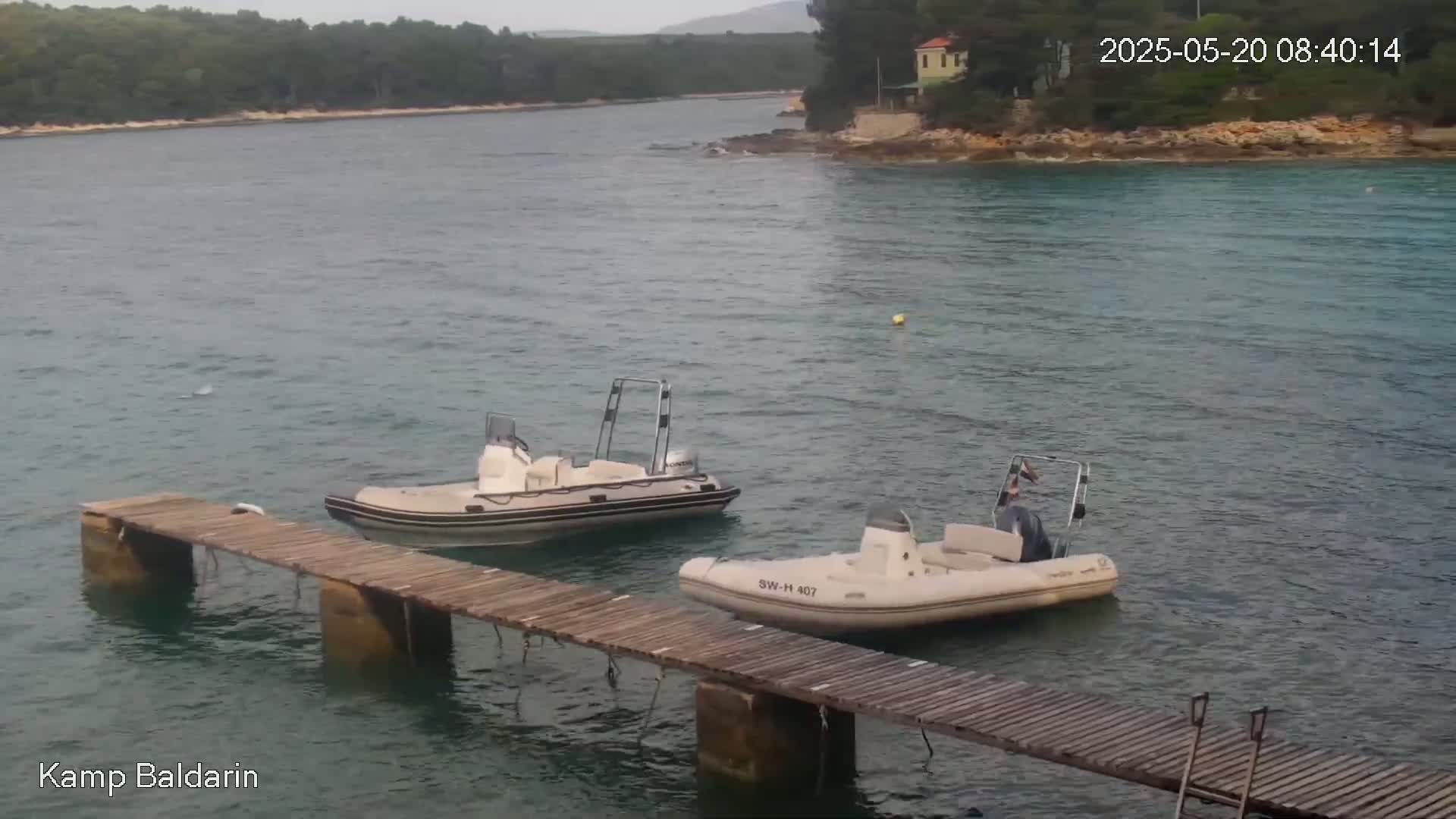 Two inflatable boats are moored near a wooden dock in calm, overcast water, with green hills visible in the background.
