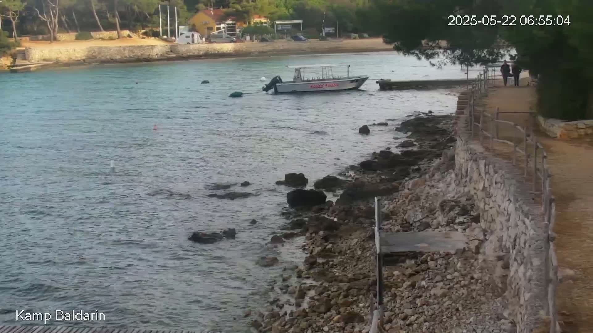 A calm body of water laps a rocky shore where a small boat is moored near a stone path with two people walking on it.