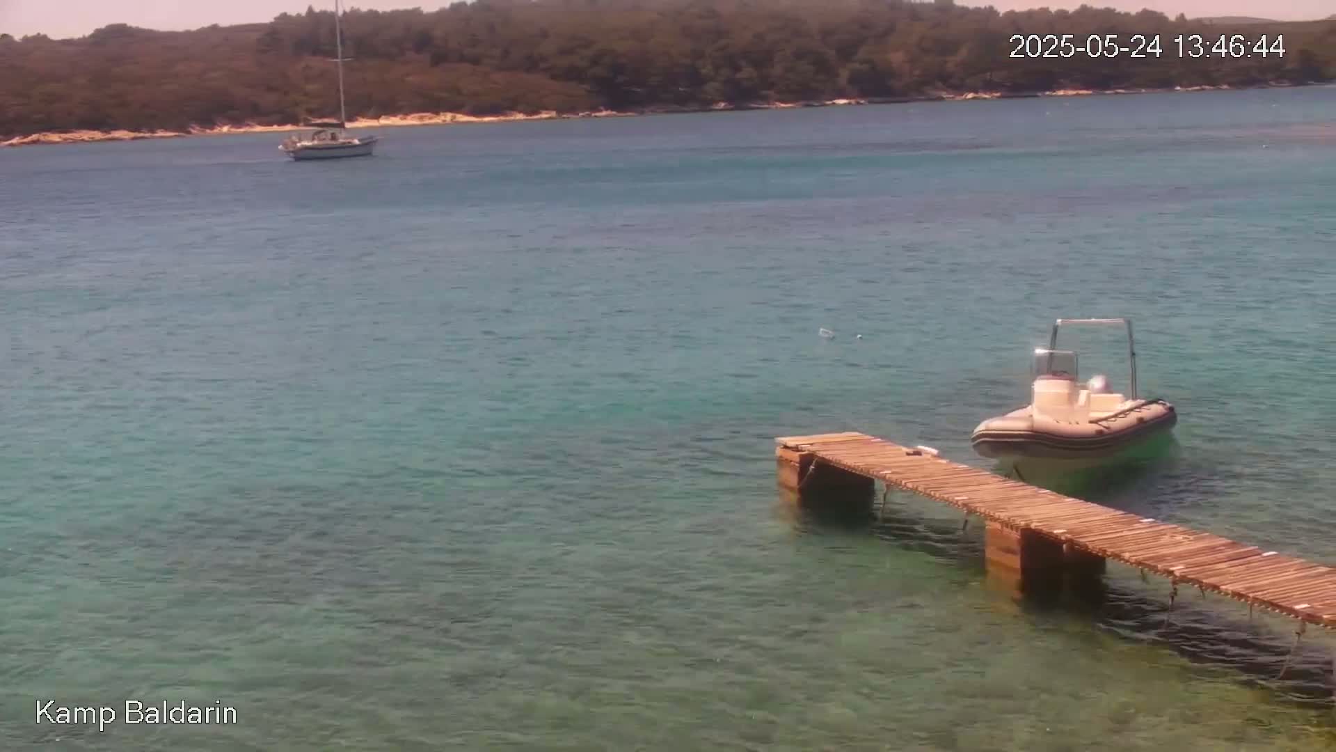 A small motorboat is moored at a wooden dock in clear, calm water near a sailboat and a wooded shore under sunny skies.