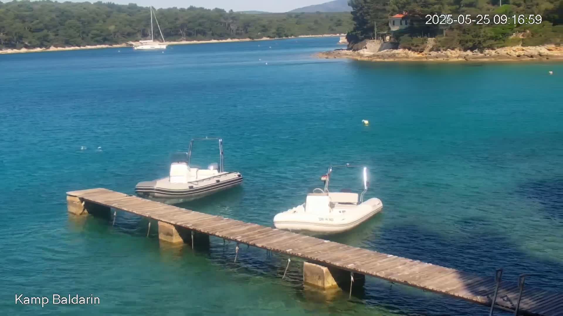Two inflatable boats are moored at a wooden dock in a calm, clear turquoise bay, with a sailboat visible in the distance and green hills in the background.