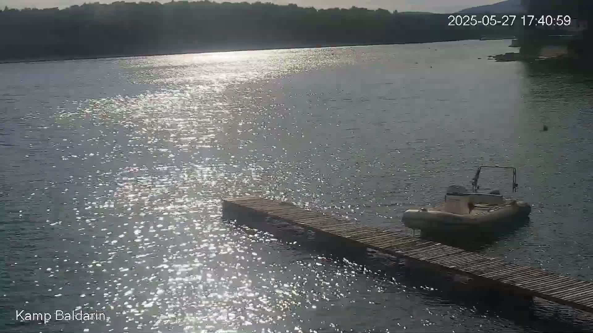 A small inflatable boat is moored at a wooden dock on a calm body of water under a sunny sky.