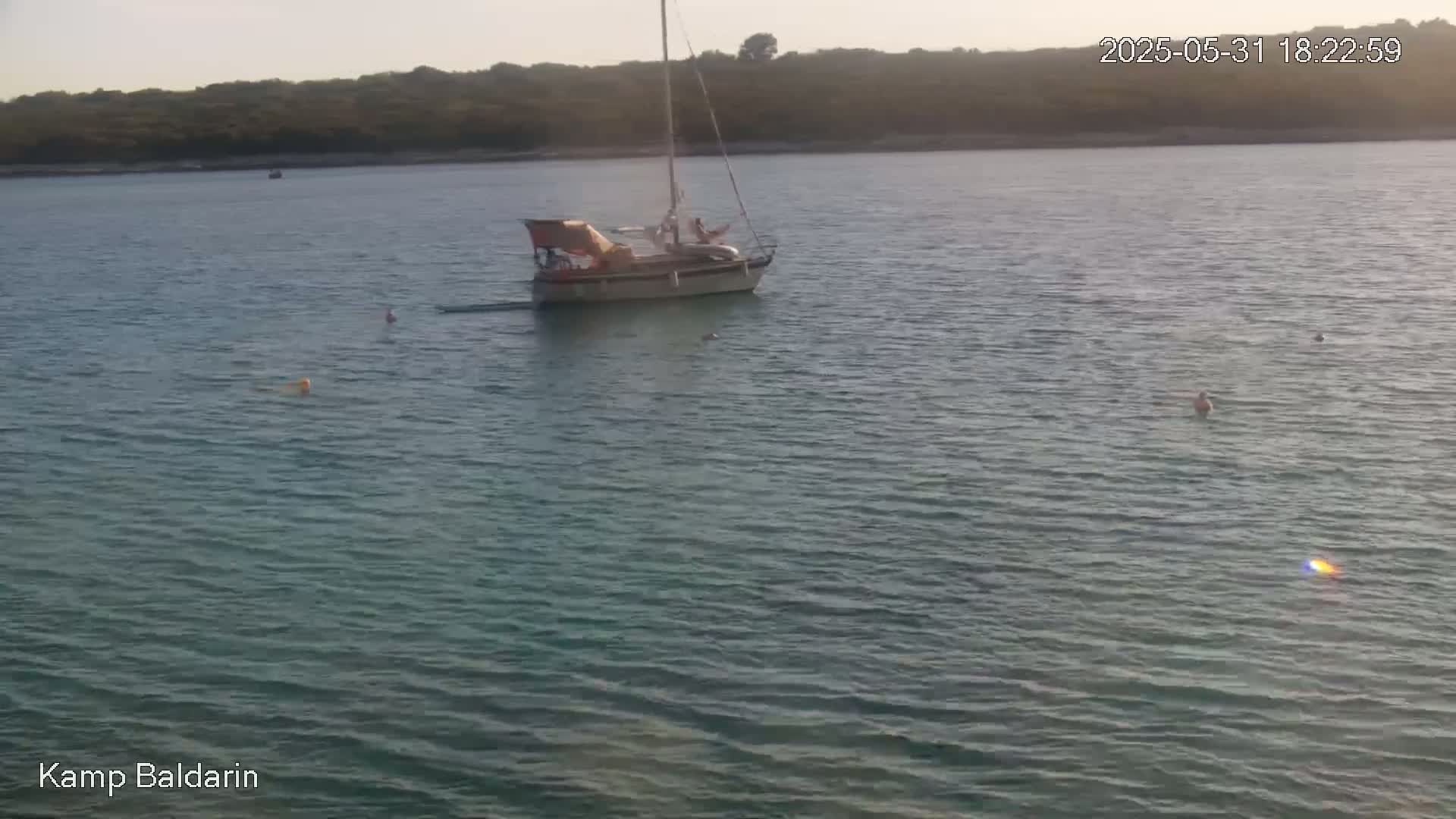 A sailboat with people on board is anchored in calm, greenish water near a shoreline of low-lying vegetation under a clear sky.