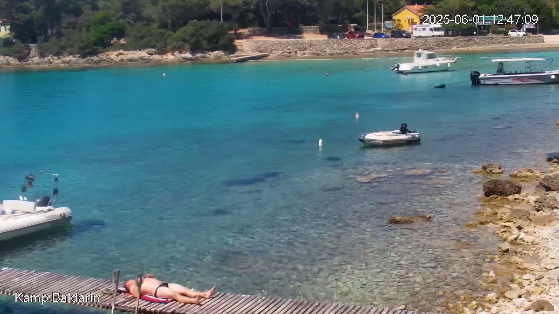 A man sunbathes on a wooden dock overlooking a calm, clear turquoise bay with several boats anchored nearby, under a sunny sky.