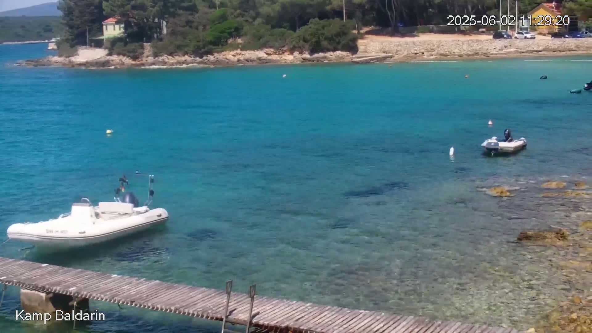 A wooden pier extends over clear, turquoise water where two small motorboats are anchored near a rocky shoreline with green trees and a small building in the background on a sunny day.