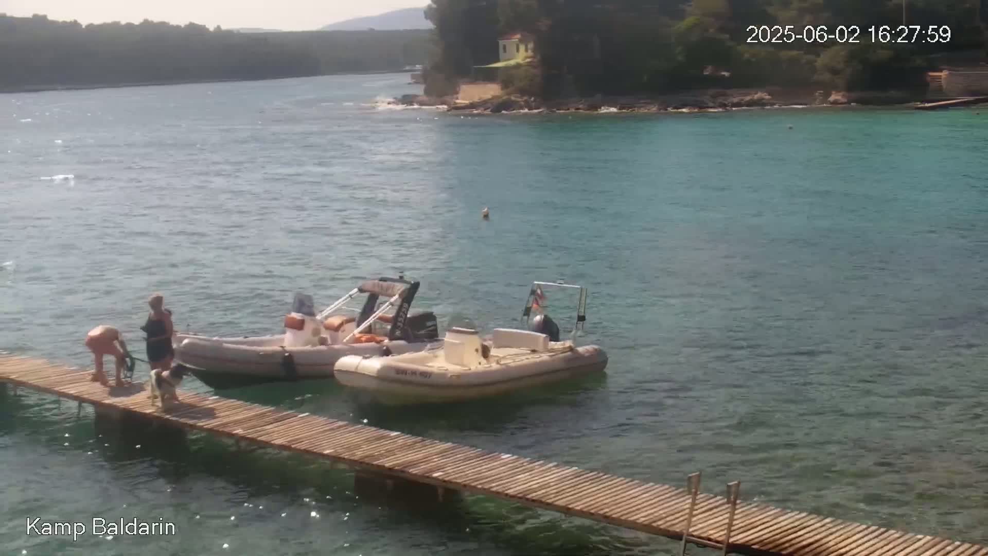 Two inflatable motorboats are moored at a wooden dock next to calm, clear turquoise water, where two people and a dog are standing.