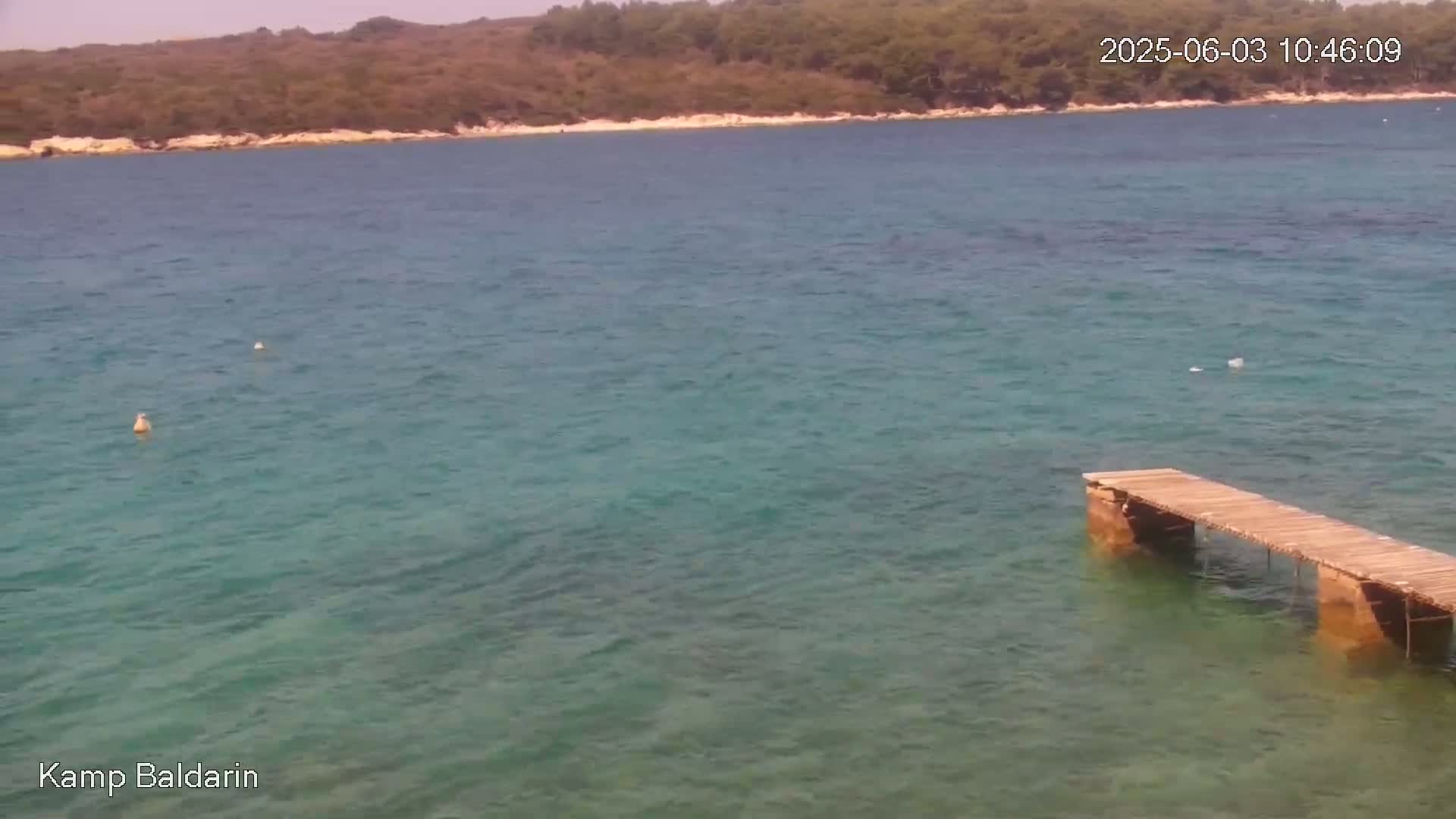 A calm, clear teal sea laps a small wooden pier extending from a rocky shoreline backed by green vegetation under a sunny sky.