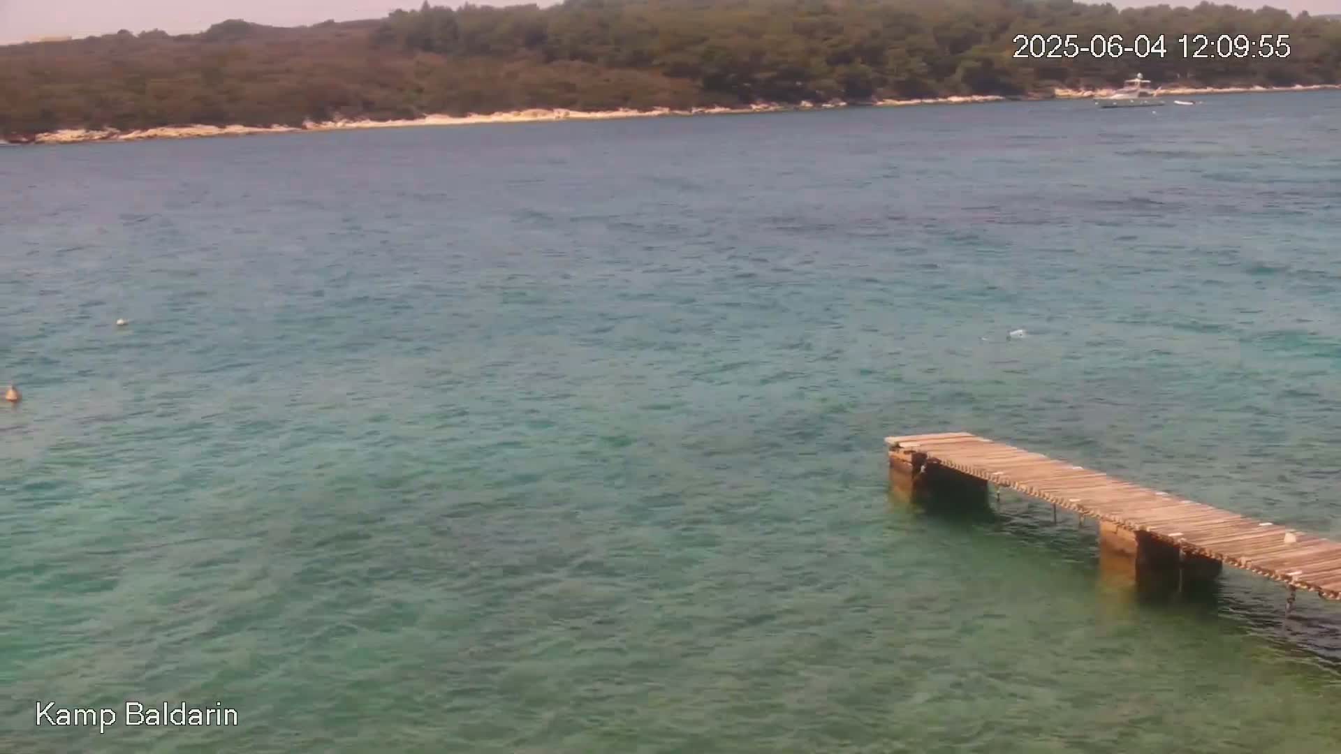 A wooden pier extends into a calm, clear teal sea, with a treeline visible in the background on a sunny day.
