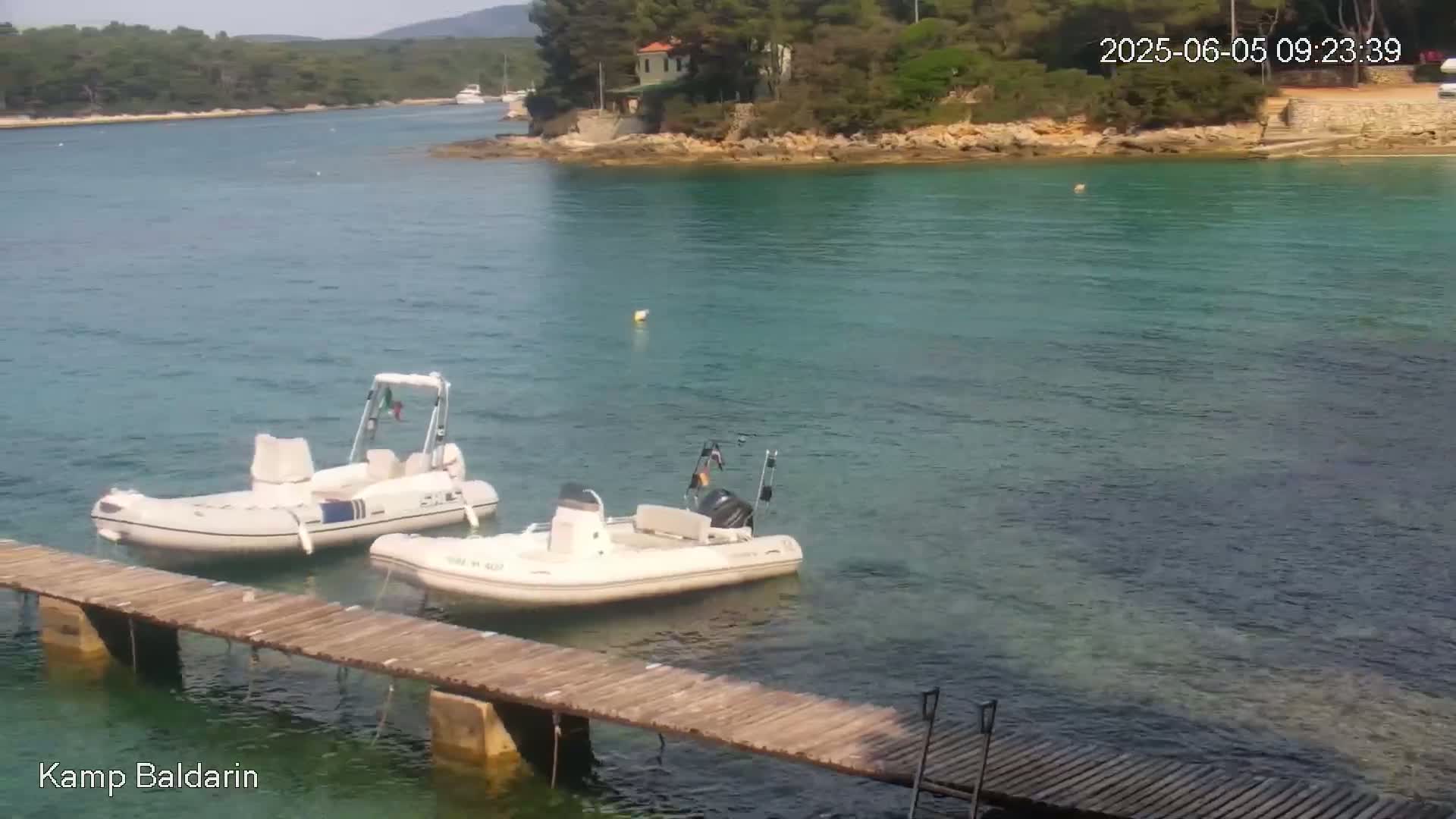 Two inflatable boats are moored at a wooden dock in calm, clear turquoise water near a verdant, rocky shoreline.