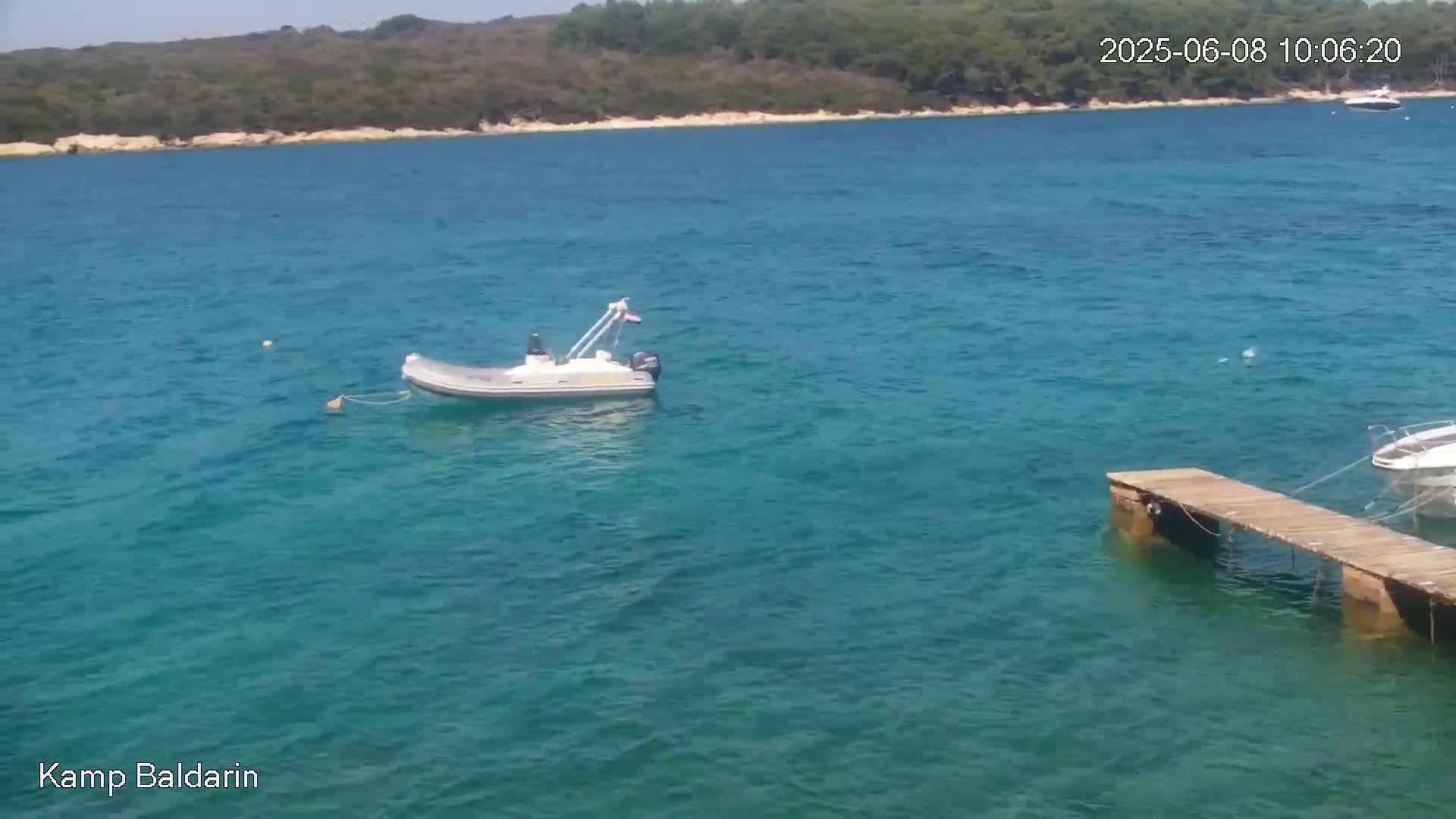 A small motorboat is moored in clear, turquoise water near a wooden pier, with a wooded shoreline in the background under a sunny sky.