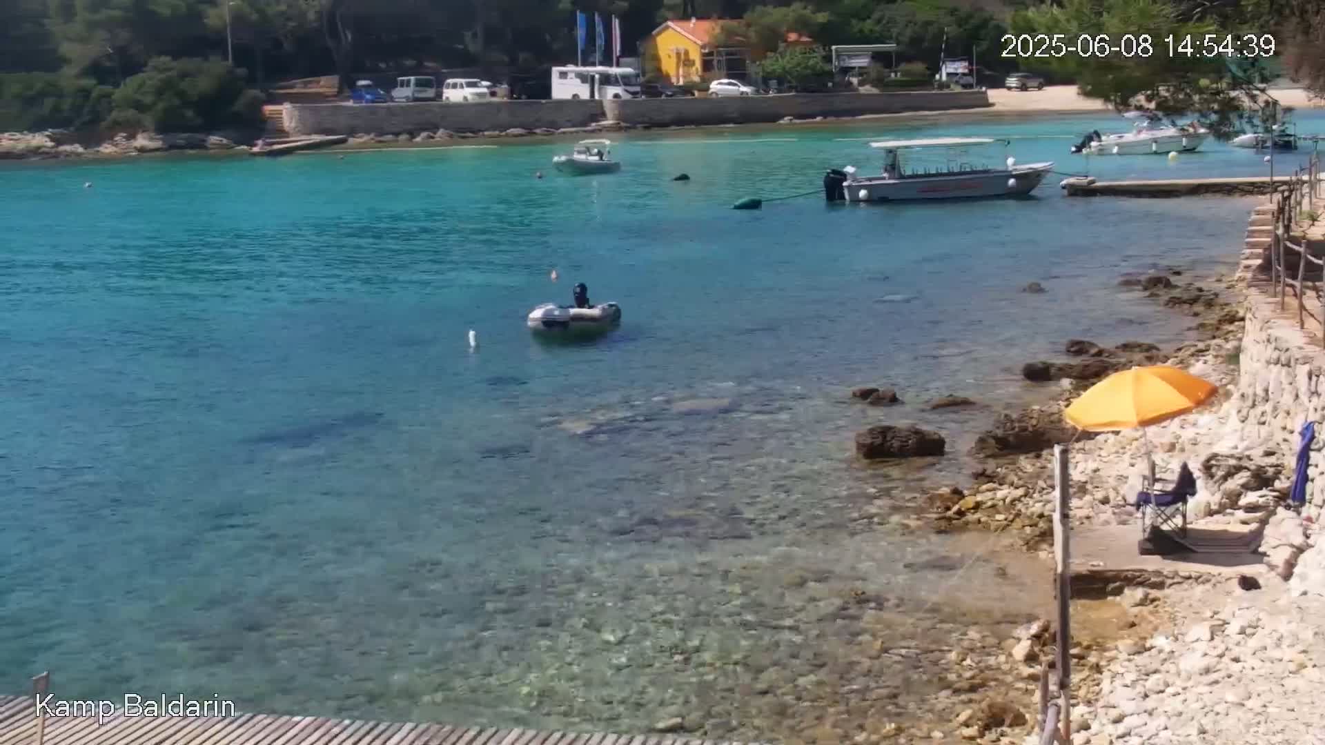 A rocky shoreline with clear, turquoise water holds several small boats, one of which is occupied, and an orange umbrella sits on a small beach next to a folding chair.
