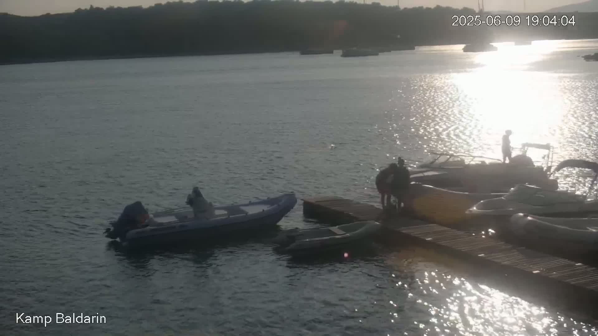 Several small motorboats are moored at a dock on calm water reflecting sunlight, with a few people standing near the boats in the late afternoon.