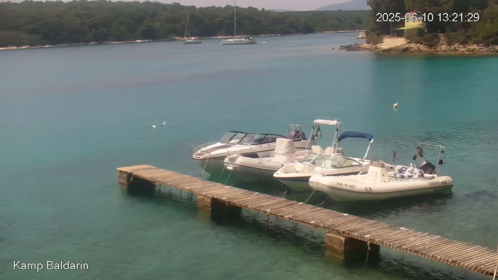Three motorboats are moored at a wooden pier in a calm, clear turquoise bay, with green hills visible in the background under a sunny sky.