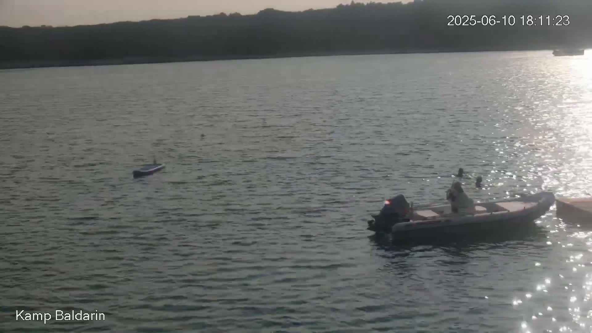 A small motorboat with people on board is on a calm body of water near a dark-colored, long, narrow object floating nearby, with a distant shoreline visible under a dusky sky.