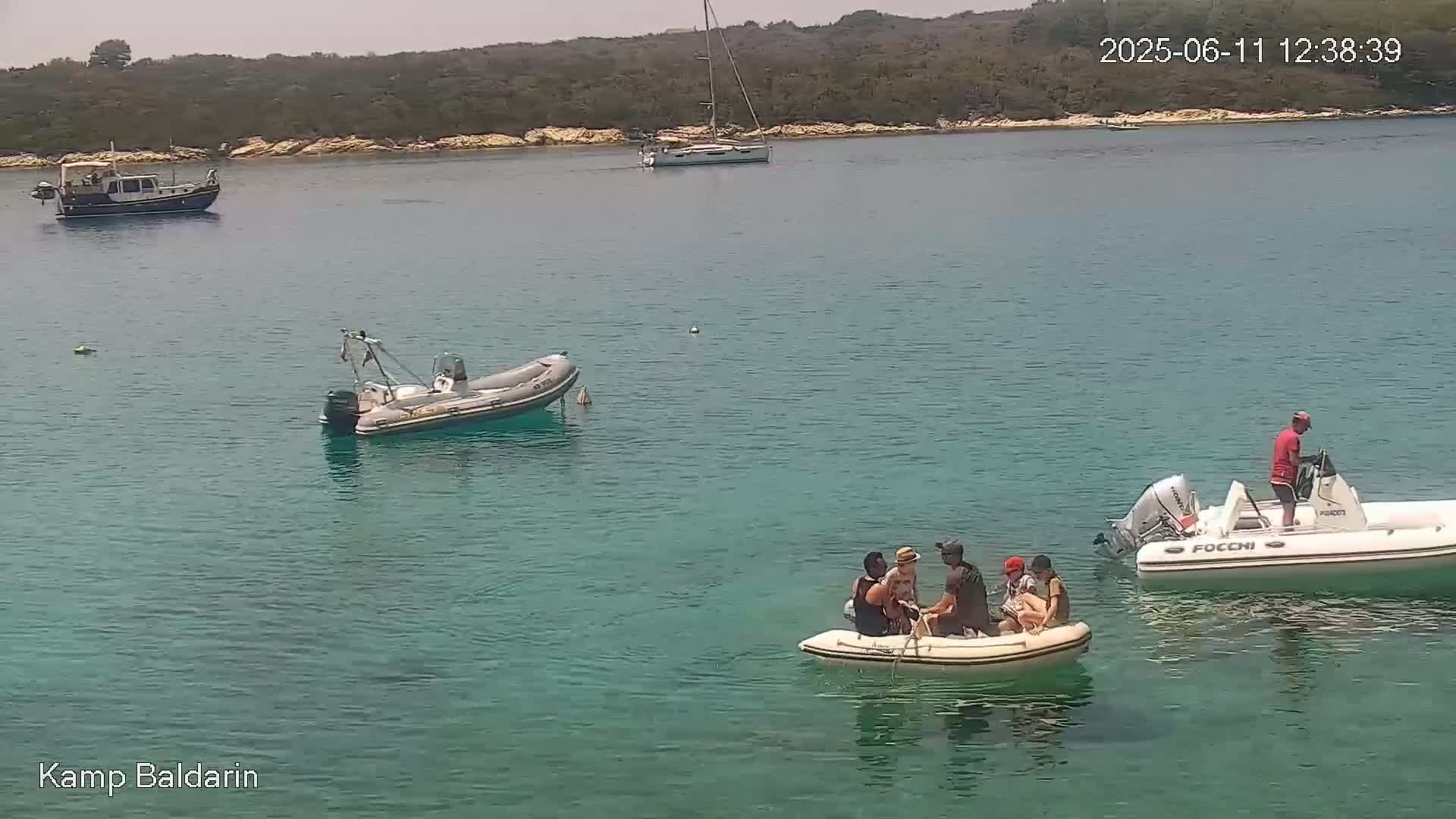 A calm, sunny day on a clear turquoise body of water shows several small boats, including a larger inflatable boat with people aboard, near a wooded shoreline.