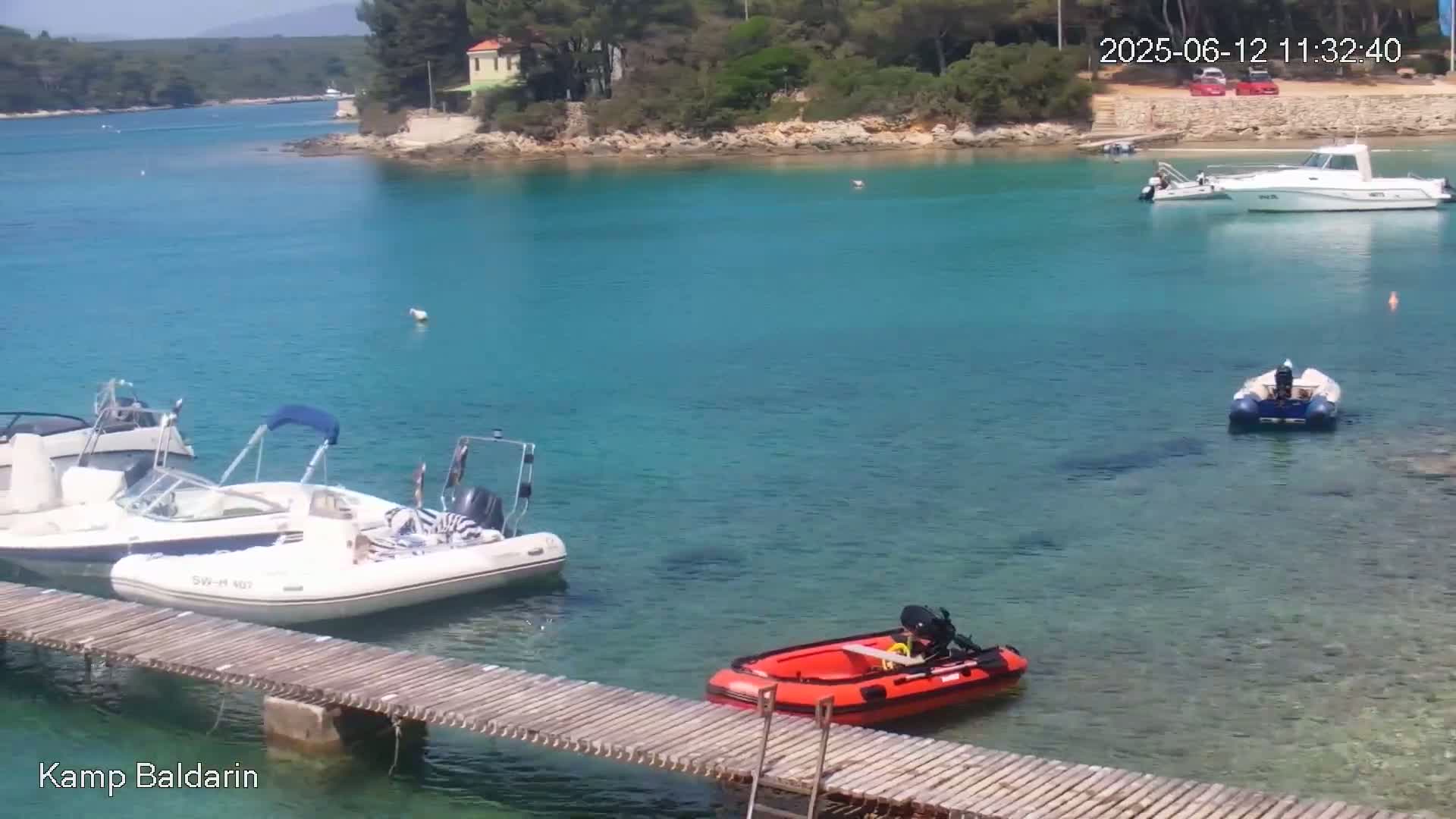 Several boats are moored in a calm, clear turquoise bay near a small, wooded peninsula on a sunny day.