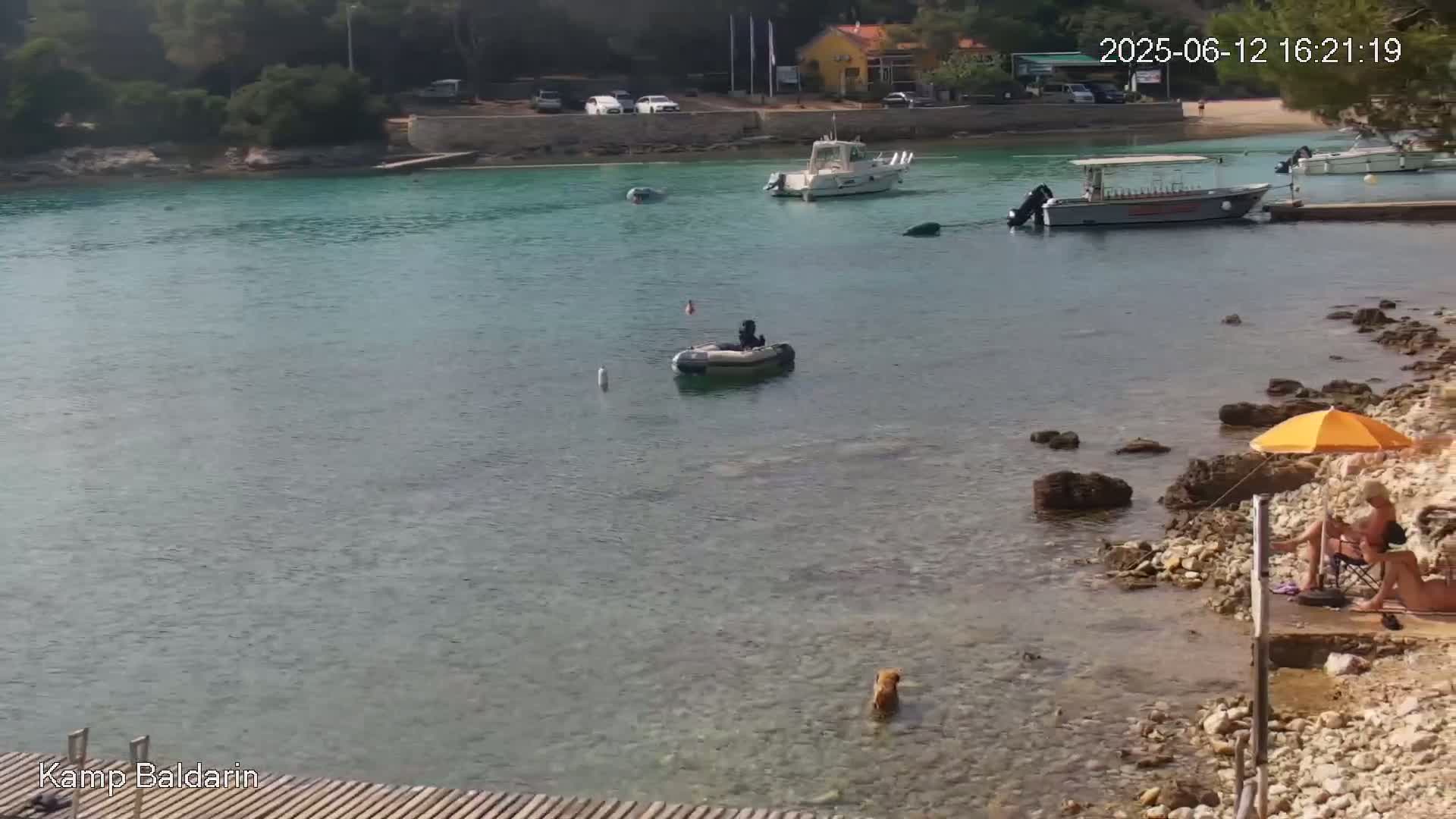 A calm, clear turquoise body of water with several boats, a small inflatable boat with a person in it, a dog in the shallows, and two people relaxing under a yellow umbrella on a rocky shore.