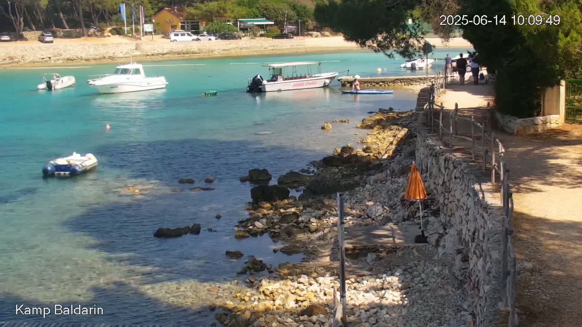 A rocky shoreline with clear turquoise water features several boats, including a small inflatable boat and a larger passenger boat, with a few people walking along a path beside a low stone wall on a sunny day.