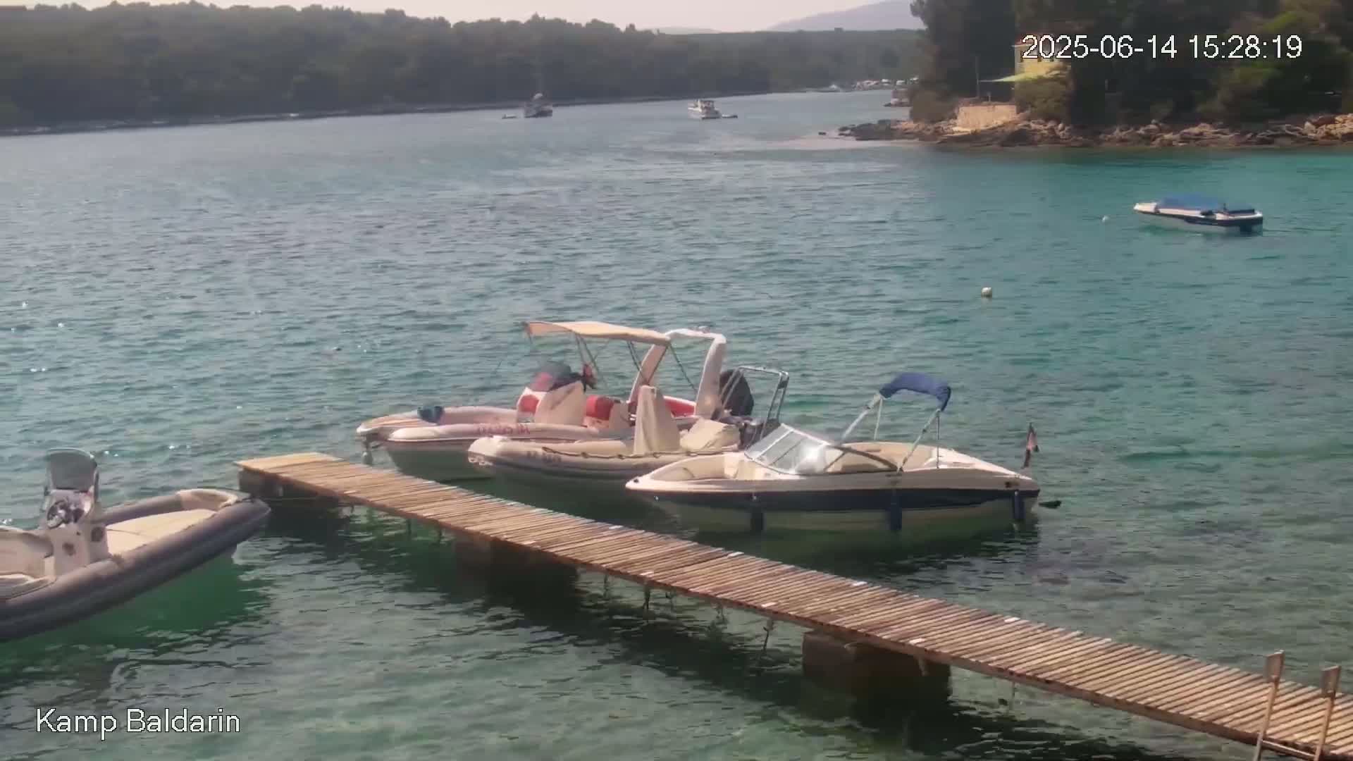 Several motorboats are moored at a wooden dock in calm, clear turquoise water near a rocky shoreline and green hills under a sunny sky.