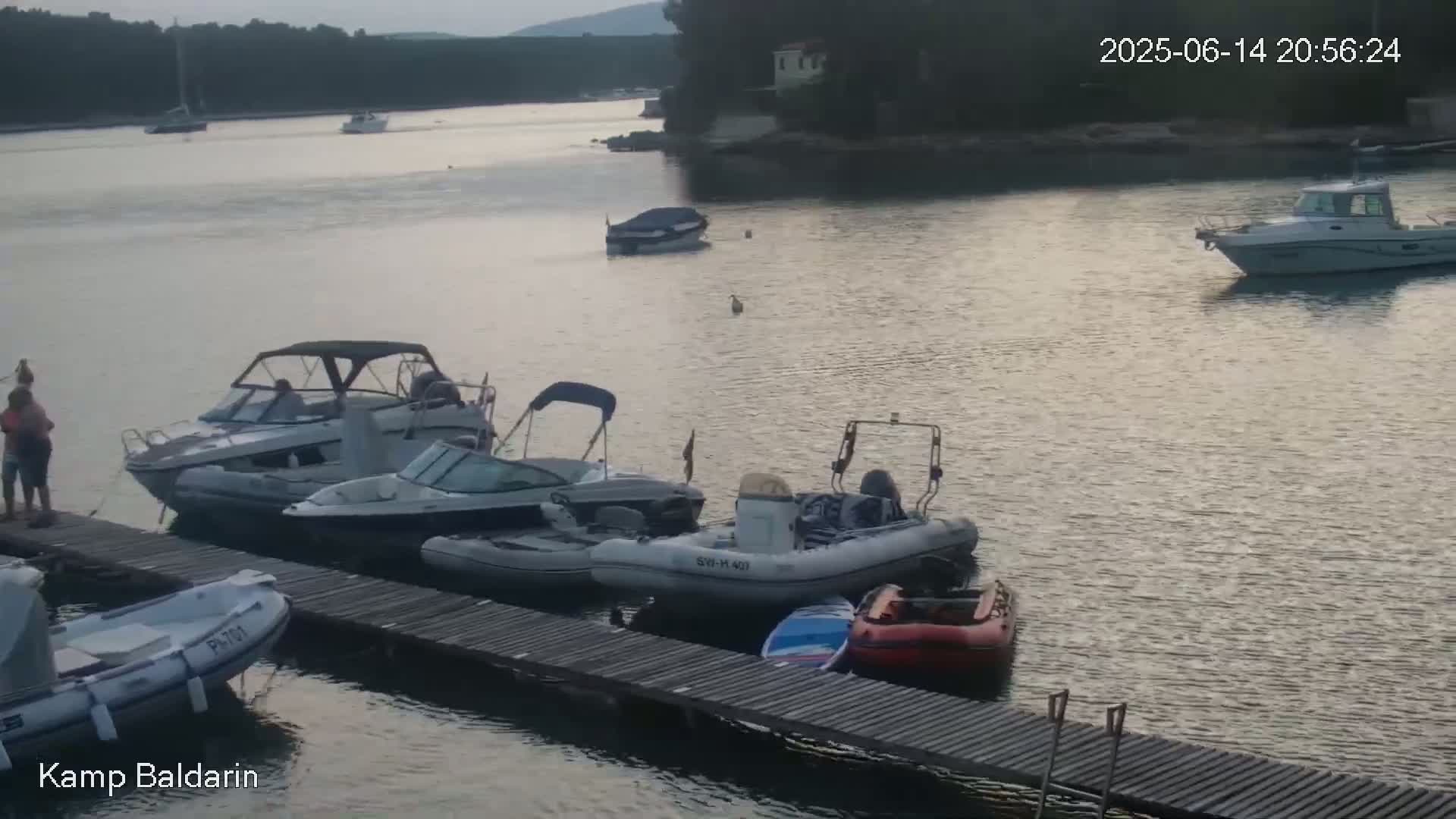 Several motorboats are moored at a wooden dock in calm, clear turquoise water near a rocky shoreline and green hills under a sunny sky.