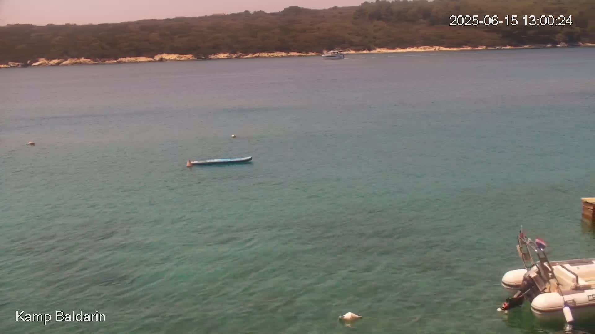 A calm, clear, turquoise body of water under a sunny sky shows a small, distant boat, a larger boat in the foreground near a dock, and a lone paddleboard.