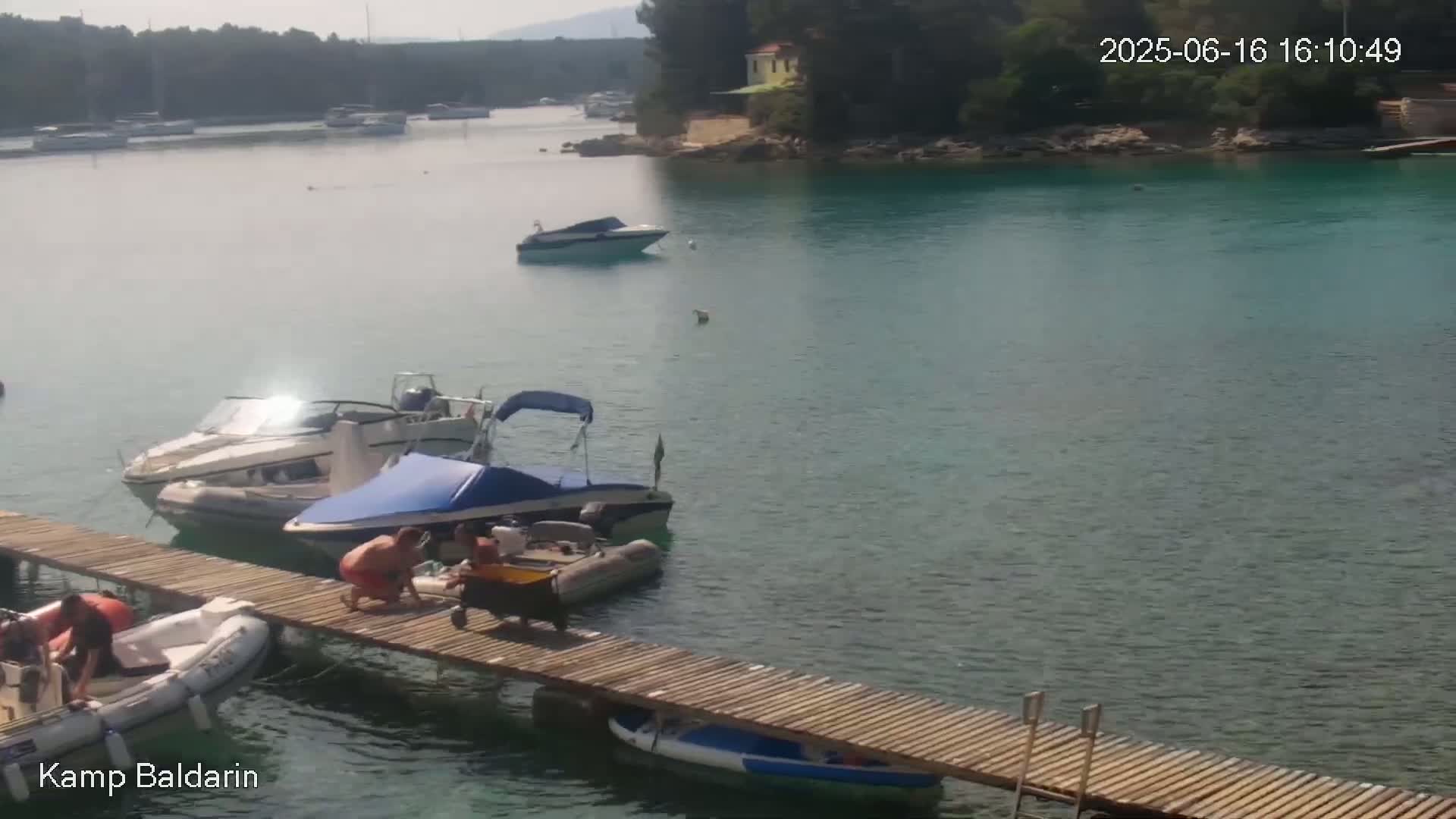 Several boats are moored at a wooden dock in a calm, clear body of water, with a few more boats visible in the distance on a sunny day.