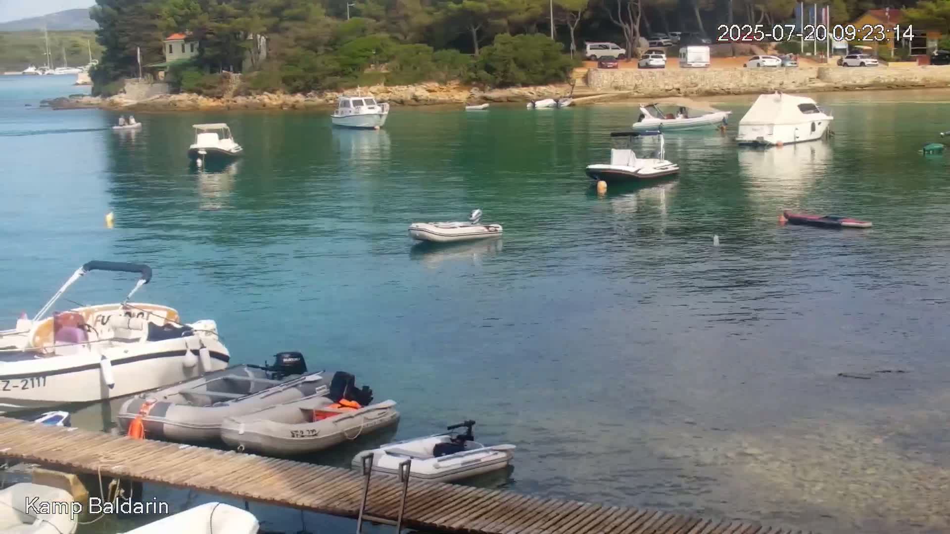 Several boats are moored in a calm, clear, teal-colored bay next to a small, wooded shoreline on a sunny day.