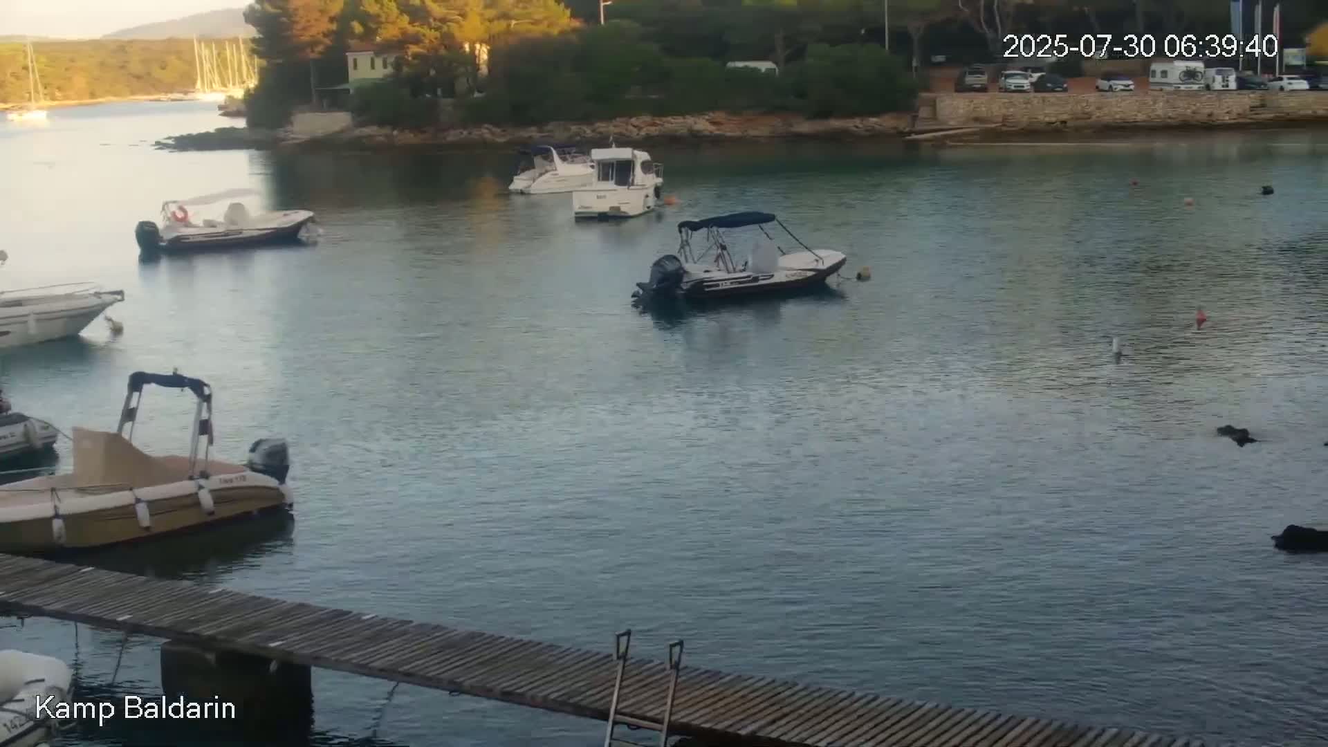 Several boats are moored in a calm, clear, teal-colored bay next to a small, wooded shoreline on a sunny day.