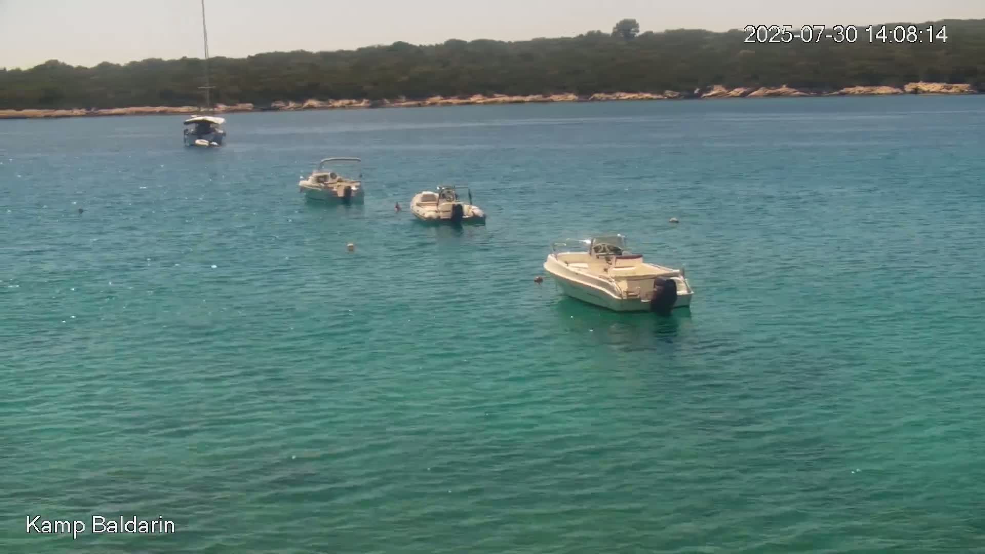 Several boats are moored in a calm, clear, teal-colored bay next to a small, wooded shoreline on a sunny day.