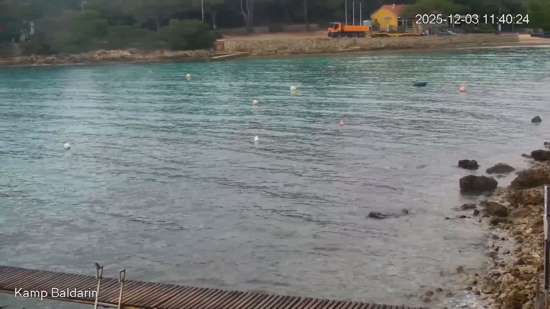 An overcast bay features several buoys on its turquoise and grey waters, with a wooden pier and rocky shore in the foreground and a distant shore showcasing a yellow building and an orange truck.