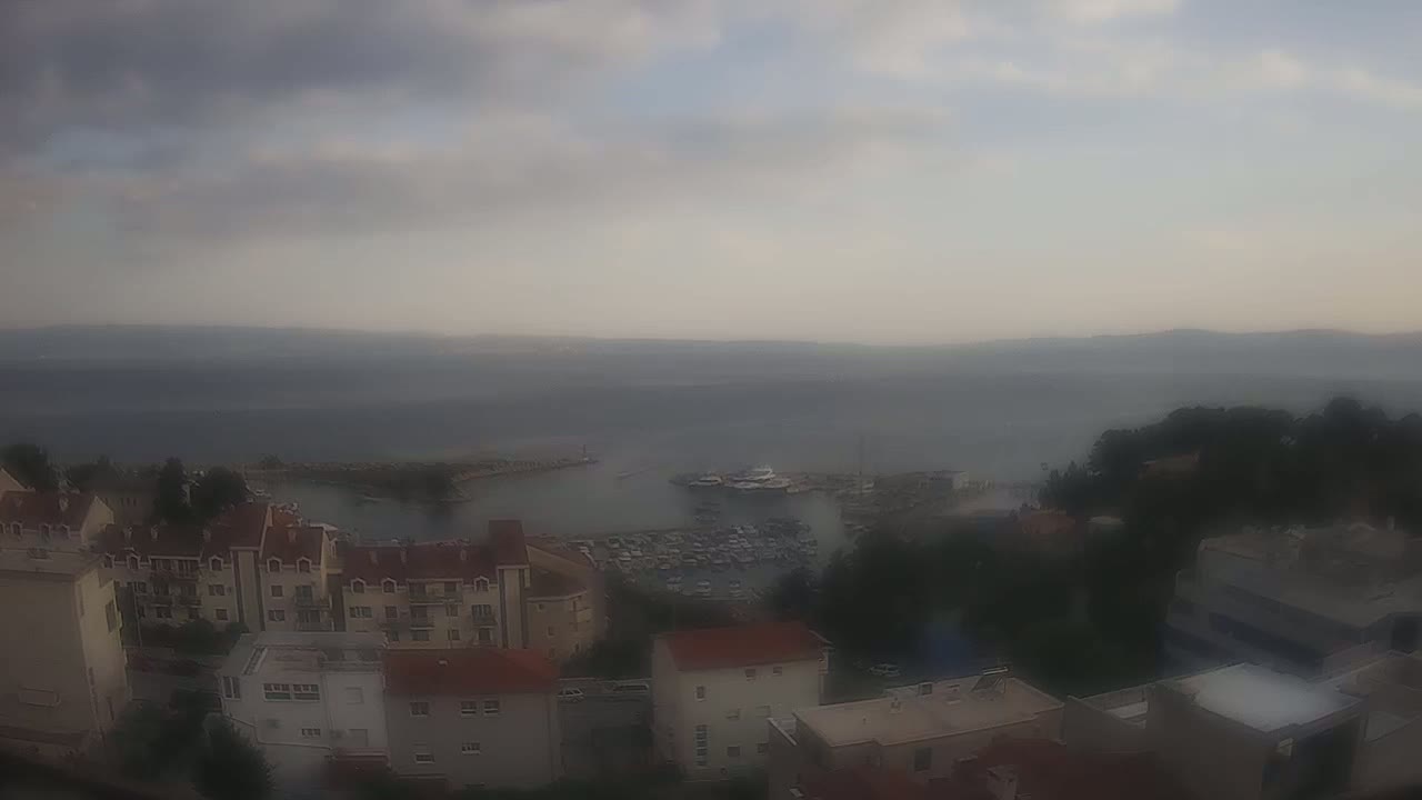 An overcast day reveals a coastal town with buildings and a marina filled with boats, overlooking a calm sea.