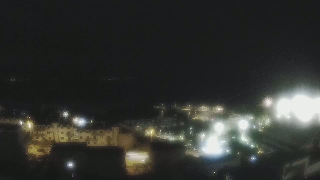 A nighttime, high-angle view shows a coastal town with buildings and illuminated areas, possibly a fireworks display in the distance.