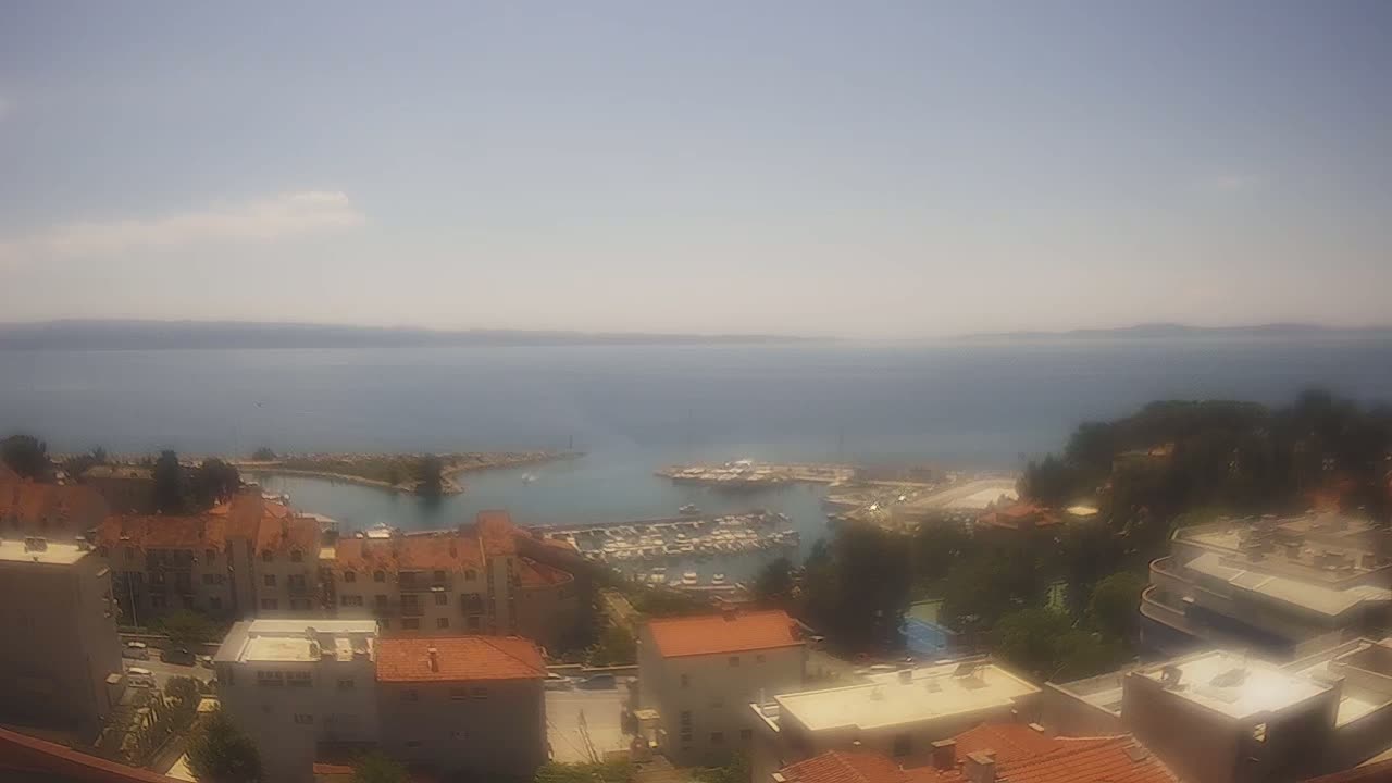 An elevated view shows a marina filled with boats, adjacent to a town of low-rise buildings under a hazy, sunny sky.
