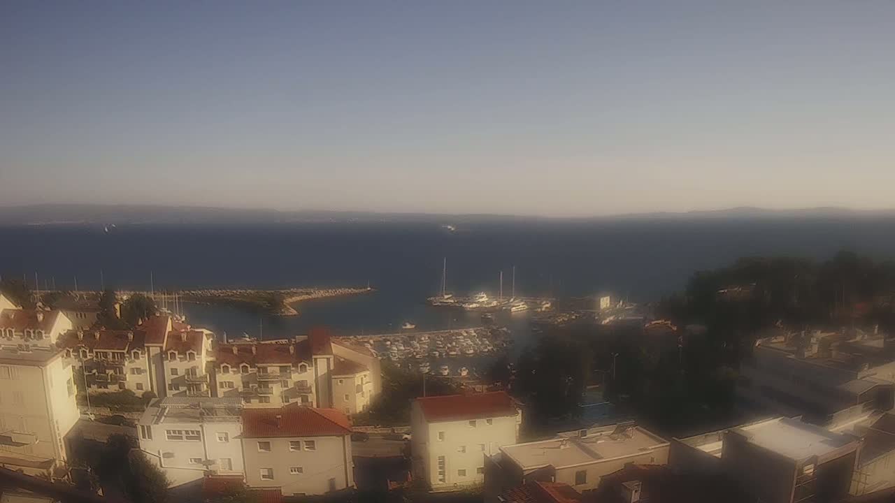 A coastal town with red-roofed buildings overlooks a calm, blue sea with a marina filled with boats under a clear sky.