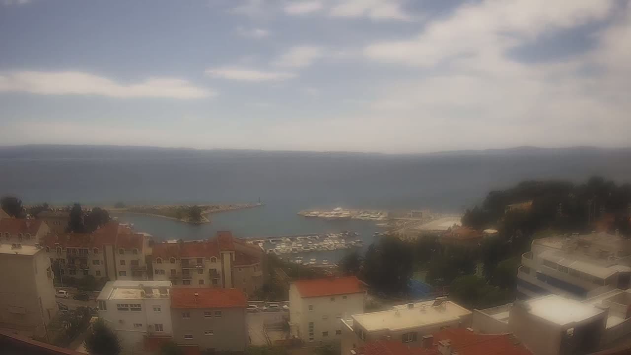 A partly cloudy day overlooks a coastal town with a marina filled with boats, and the calm sea extending to the horizon.