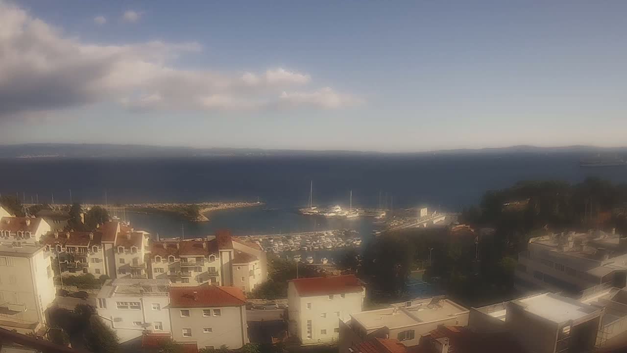 A partly cloudy day reveals a coastal town overlooking a calm harbor filled with boats, stretching to a distant coastline.