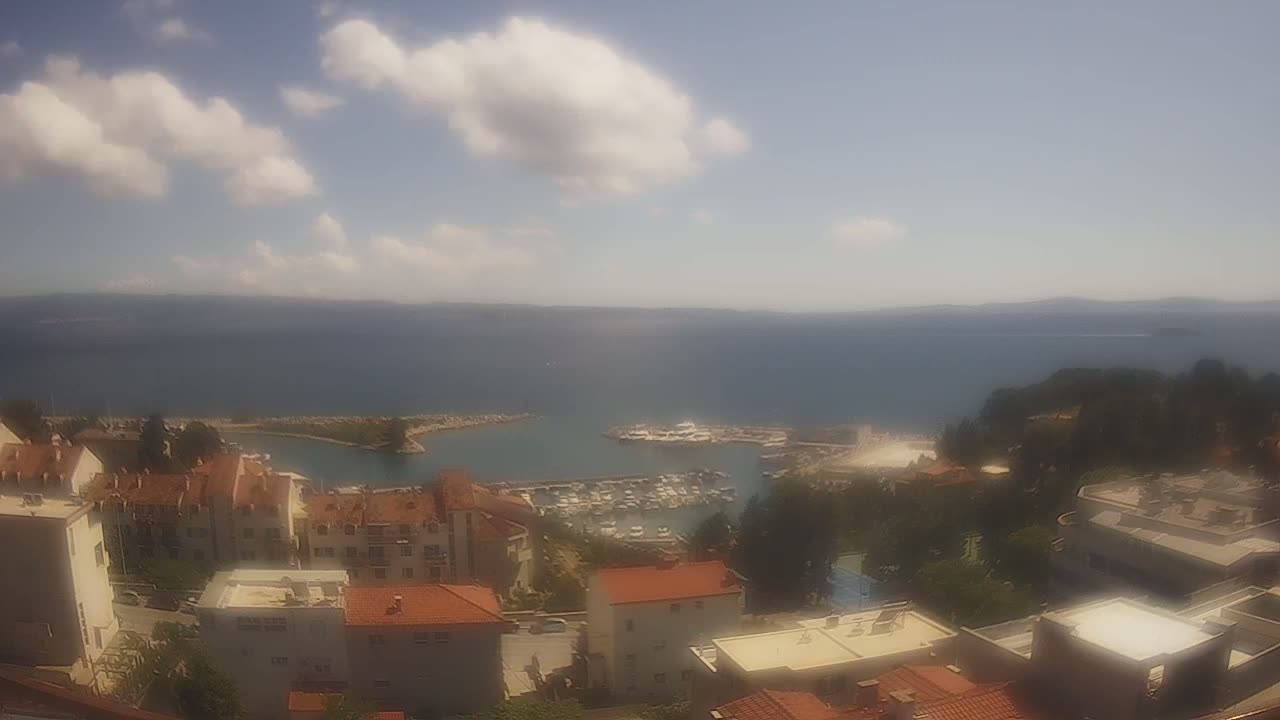 A partly cloudy day overlooks a coastal town with a marina filled with boats, and the ocean stretching to a distant horizon.