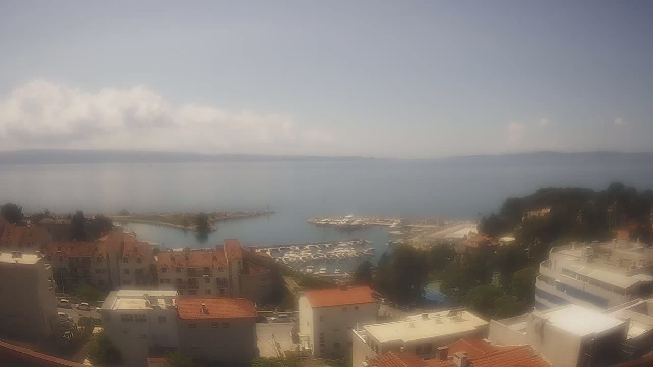 A hazy, sunny day overlooks a marina filled with boats, nestled between a hillside of buildings and a calm sea stretching to a distant shoreline.