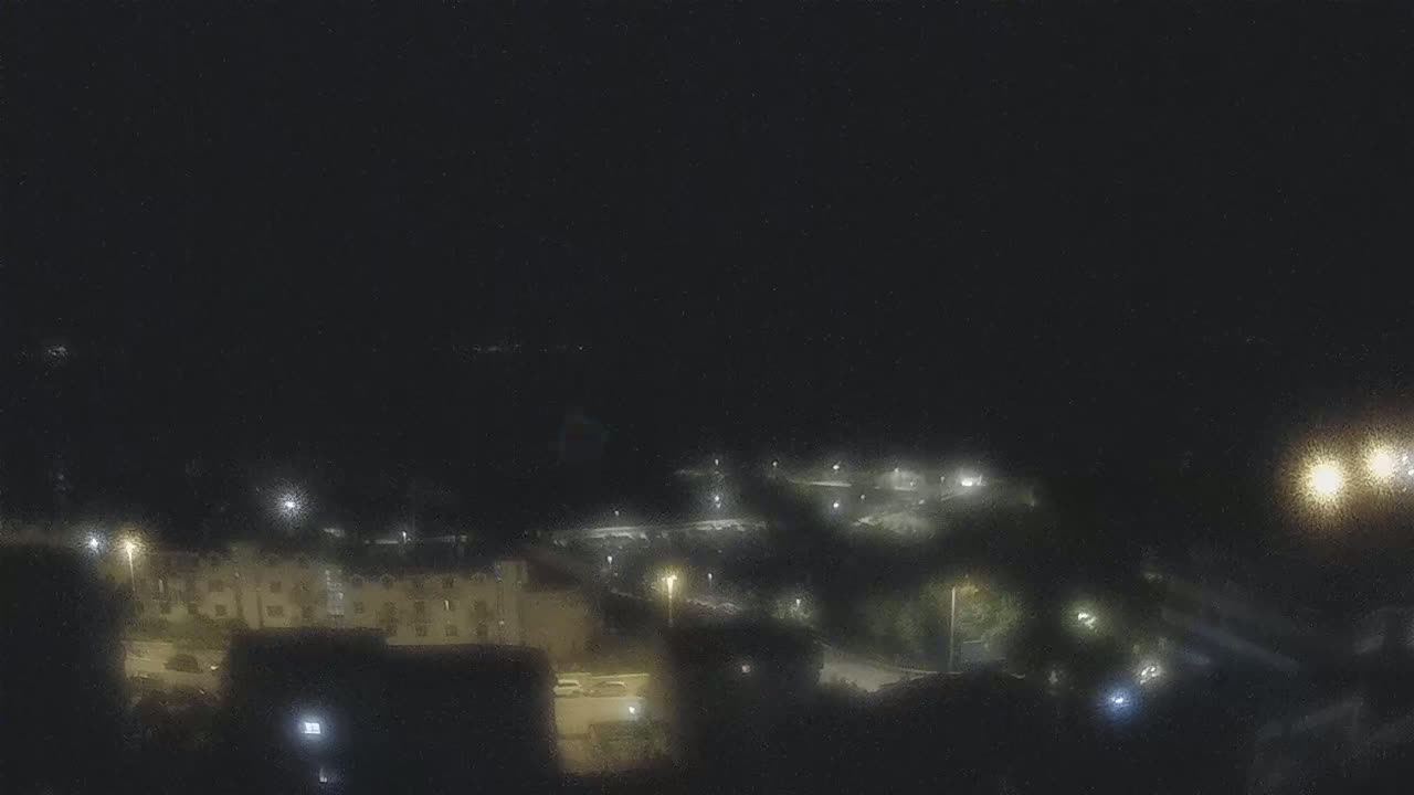 A nighttime, aerial view shows a dimly lit residential area with several buildings and streetlights under a dark sky.