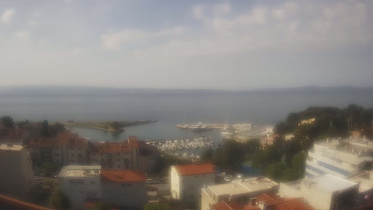 A hazy, partly sunny day overlooks a coastal town with a marina filled with boats,  buildings with red tile roofs, and a calm sea extending to the horizon.