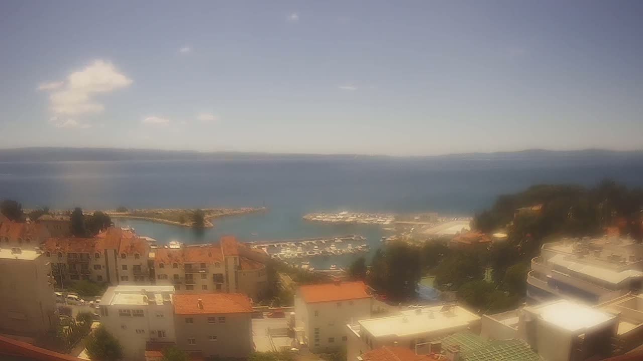 A partly sunny day overlooking a coastal town with a marina filled with boats, and a calm sea stretching to a distant shoreline.