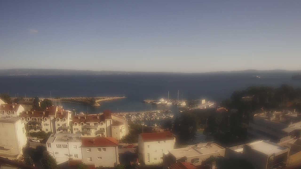 A coastal town with red-roofed buildings overlooks a calm blue sea filled with boats in a marina on a clear, sunny day.