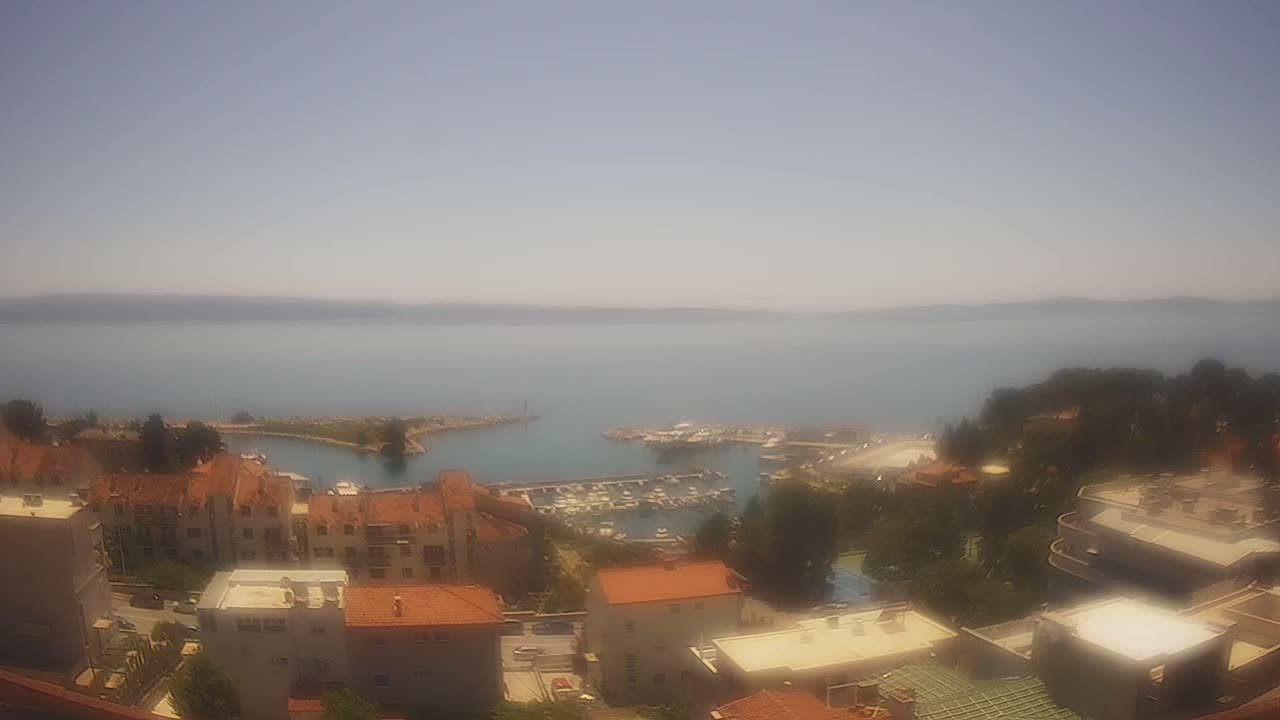 A hazy, sunny day overlooks a small marina filled with boats, nestled between a coastal town of red-tiled buildings and a calm blue sea.