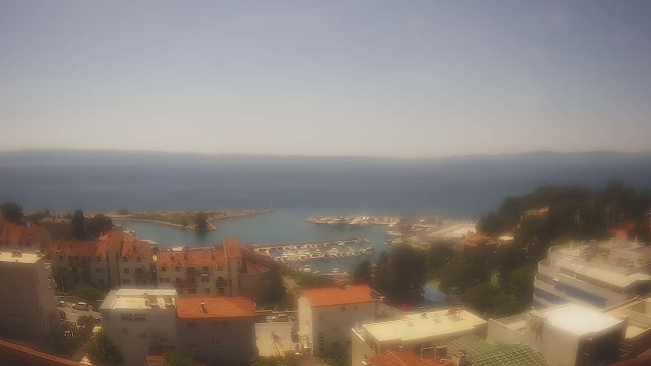 A hazy, sunny day overlooks a coastal town with a marina filled with boats, surrounded by buildings and greenery, and a calm sea extending to the horizon.