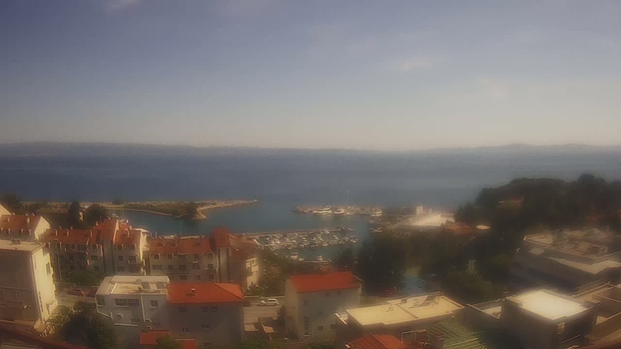 A hazy, sunny day overlooks a coastal town with red-roofed buildings clustered around a marina filled with boats, extending to a calm sea and distant mountains.