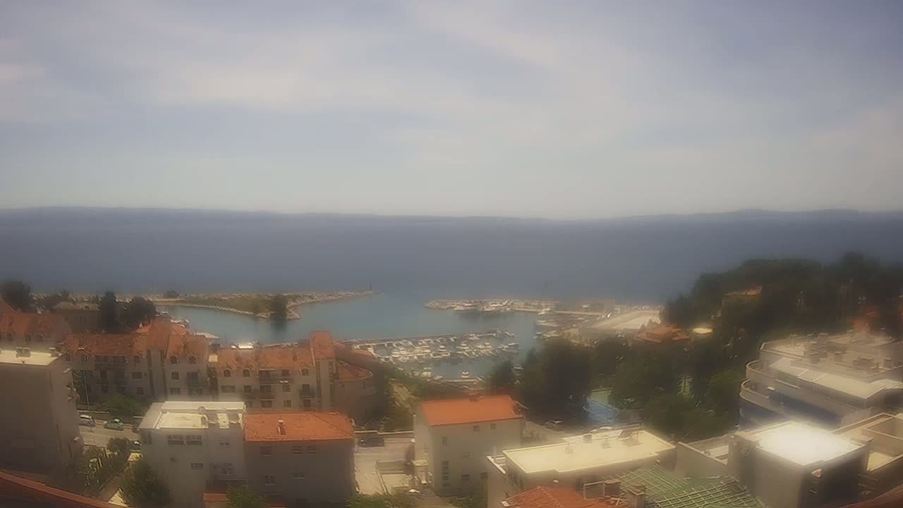 A hazy, sunny day overlooks a coastal town with a marina filled with boats, and red-roofed buildings nestled amongst green trees.