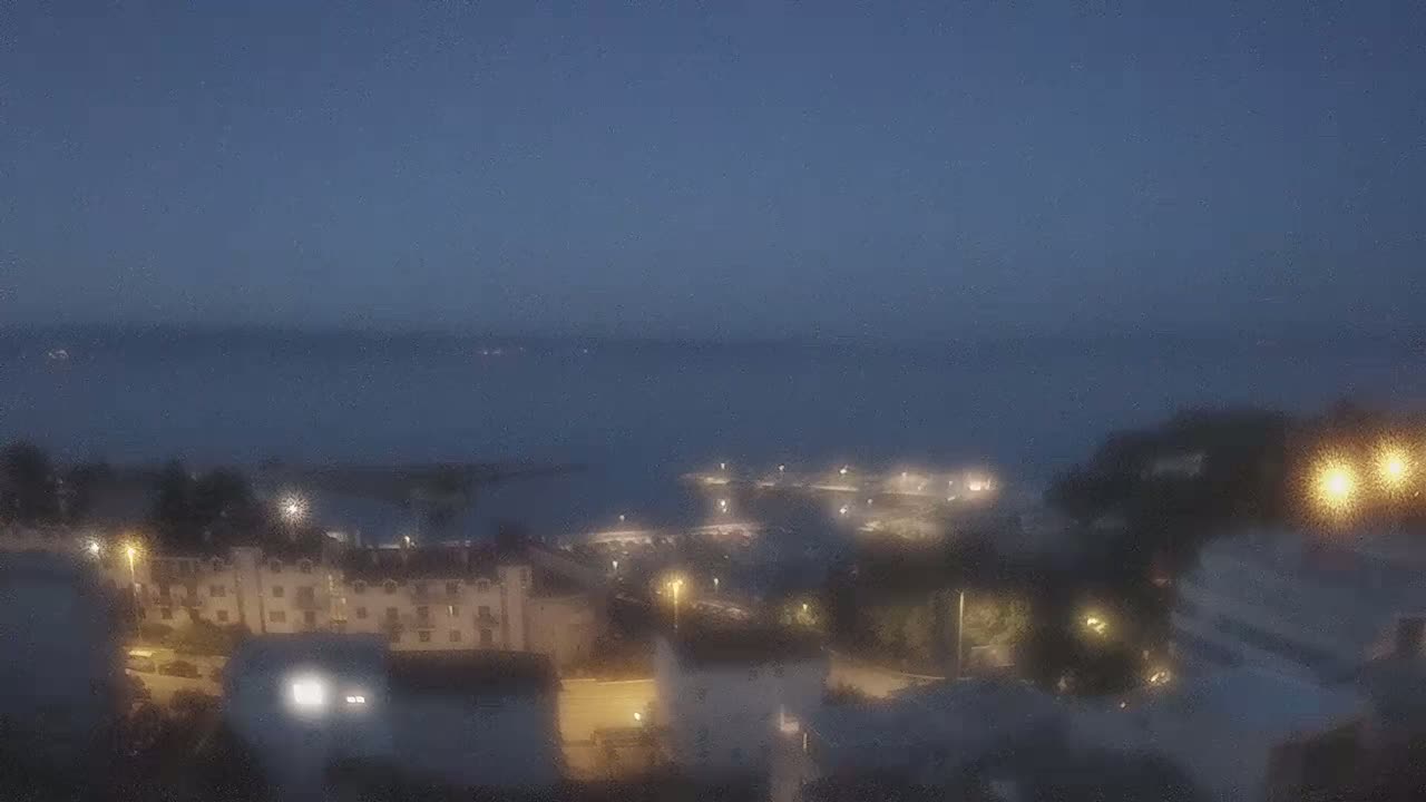 A nighttime view of a coastal town, with dimly lit buildings and a calm body of water in the background.